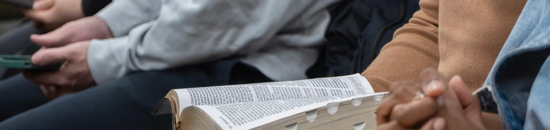 Hands resting on laps, one holding a book, others on phones, in a seated group setting.